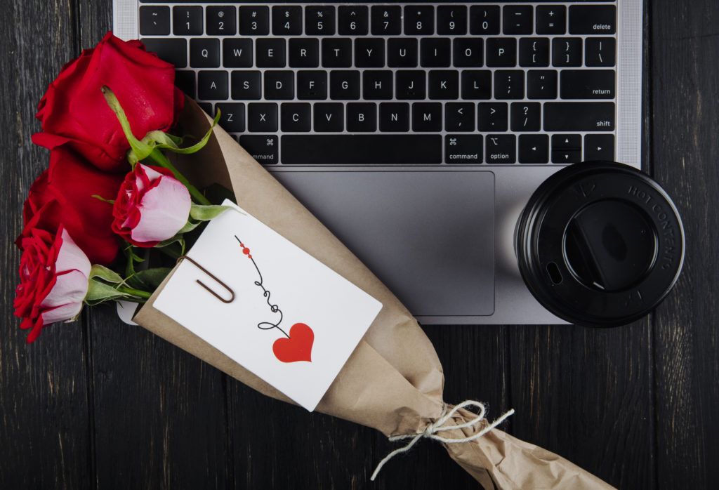 top view a bouquet of red roses in craft paper with attached postcard lying on a laptop with a paper cup of coffee on dark wooden background